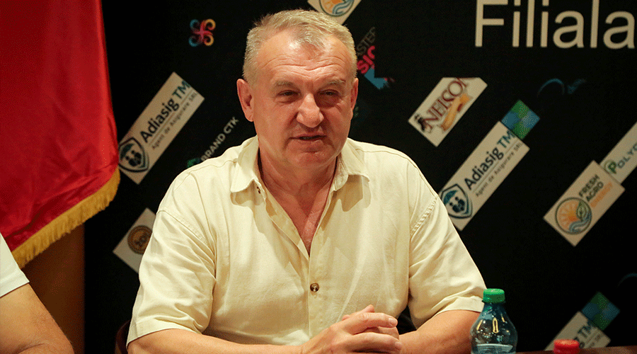 casa-de-comert-chineza-3 - Sursa de Vest Middle-aged man in a cream shirt seated at a table during a panel, with a logo-covered backdrop and a red flag nearby.