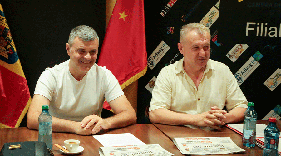 casa-de-comert-chineza - Sursa de Vest Two men at a press conference table with red flags behind them and water bottles on the desk, smiling at the camera.
