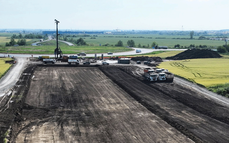 centura-vest - Sursa de Vest Construction site in a rural area with a tall drilling rig, several trucks, and dark excavated soil on a wide dirt road; fields and a curved road in the distance.