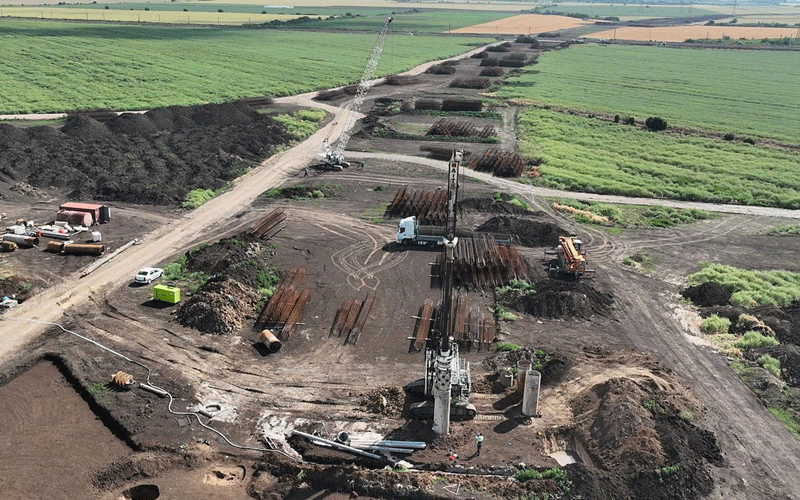 centura-vest4 - Sursa de Vest Industrial construction site with a drilling rig, piles of steel pipes, and dirt roads in a rural farmland setting, background fields visible.