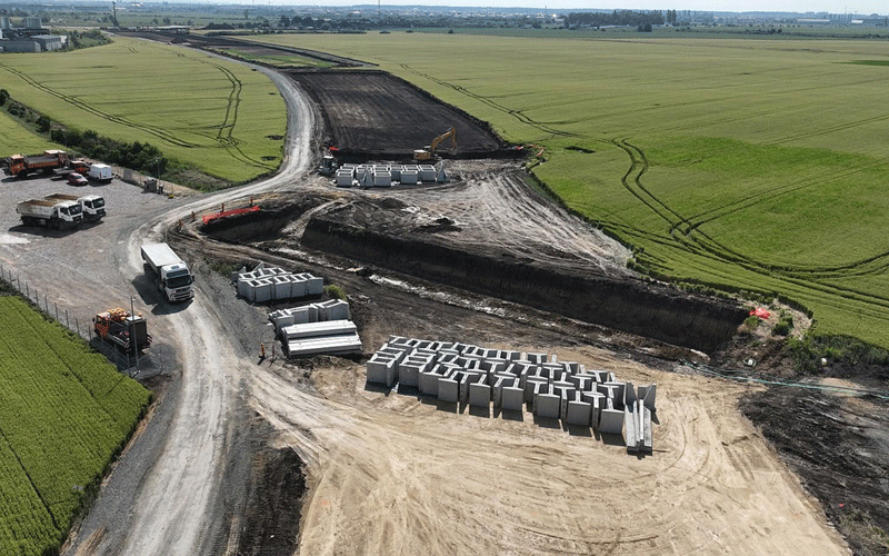 centura-vest5 - Sursa de Vest Construction site in a rural field with dirt roads, concrete barrier blocks, and a yellow excavator moving earth.
