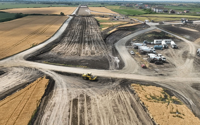 Aerial view of a large excavation site in farmland, with curved dirt paths, heavy machinery, and storage trailers to the right.
