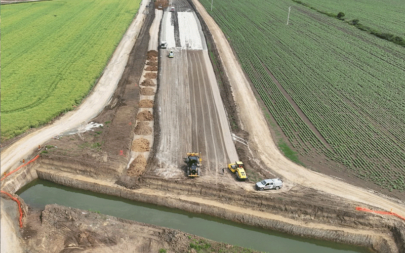 centura-vest8 - Sursa de Vest Aerial view of a road construction site between green fields, with heavy machinery spreading gravel and a concrete path under construction by dirt embankments.
