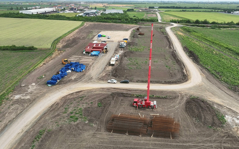 centura-vest9 - Sursa de Vest Aerial view of a rural construction site with a tall red crane, stacked steel bars, trucks, and portable buildings on dirt roads amid fields.
