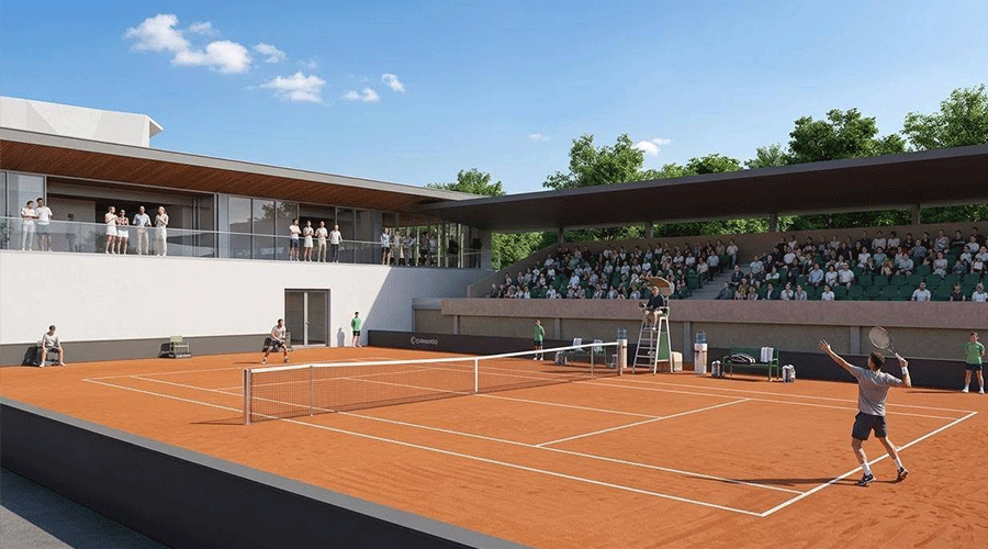 Tennis match on a clay court with players serving while a crowd watches from under a shaded stands.