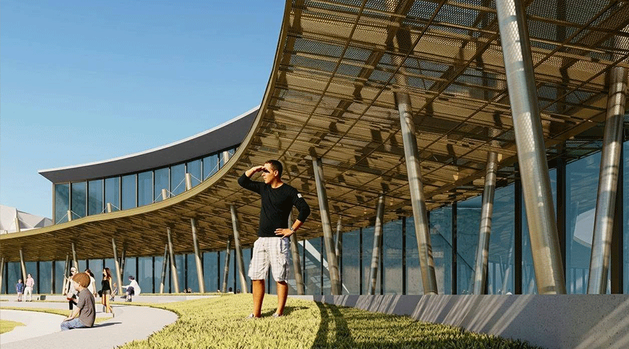 Man in a black long-sleeve shirt and white shorts stands on a grassy area outside a curved glass building, shielding his eyes from the sun while looking into the distance, with others seated nearby.