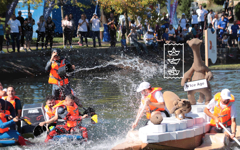 Team in orange life jackets race a raft on a sunny watercourse while spectators watch from the bank. Splashing water and a decorative float with banners are visible in the scene.