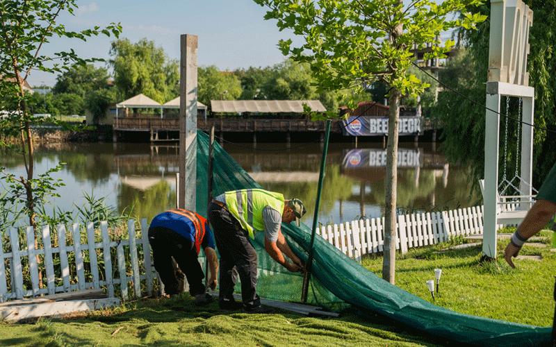 Two workers in safety vests unrolling a green net along a lakefront park with a white picket fence nearby.