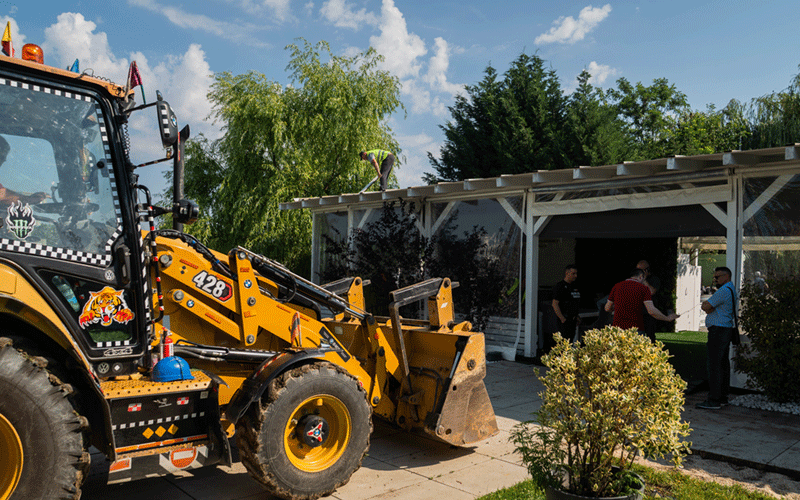 Yellow backhoe loader with front bucket parked beside a white pavilion while people stand nearby in a sunny yard.