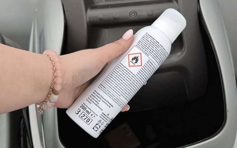 Hand holding a white aerosol spray can with a hazardous materials label (flame symbol) inside a car.