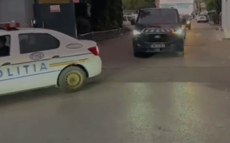 White police car with blue and yellow stripes labeled POLITIA on a city street; a dark SUV approaches with headlights on.