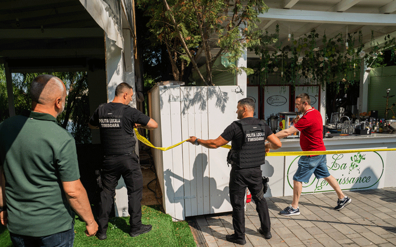 Two local police officers in black uniforms cordon off an outdoor cafe area with yellow tape as a man in a red shirt watches.