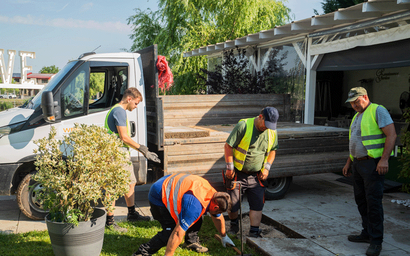 Four workers in reflective vests dig a trench beside a white van to lay a new sidewalk outside a house.