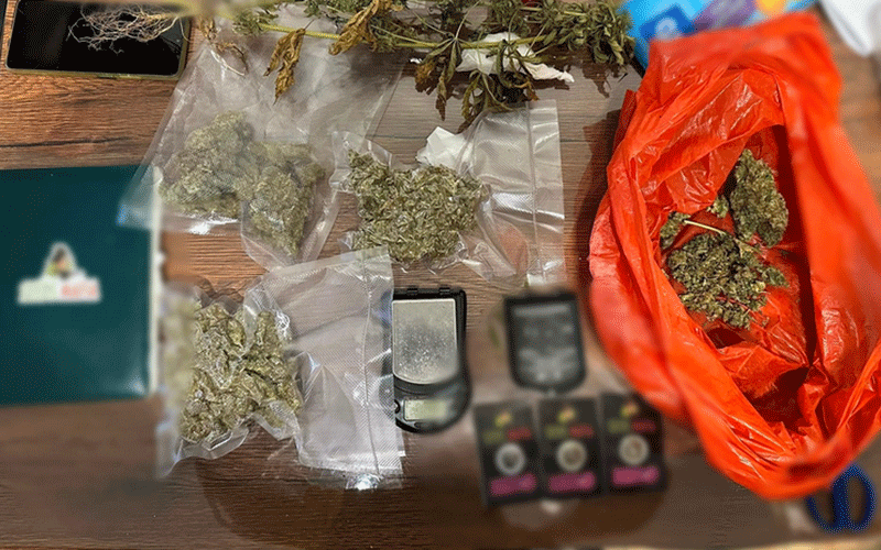 Top-down view of cannabis buds in plastic bags on a wooden table, with a red plastic bag and digital scales nearby on the surface.