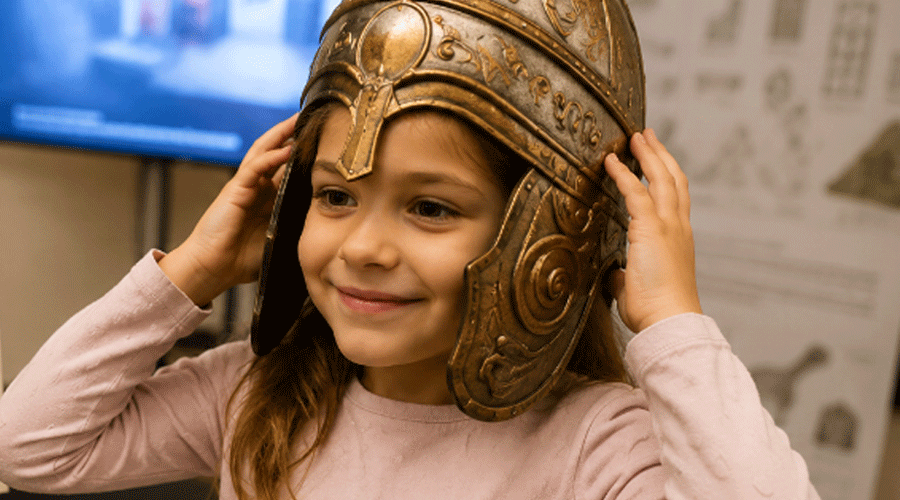 Young girl smiles while trying on a bronze helmet in a museum or classroom setting