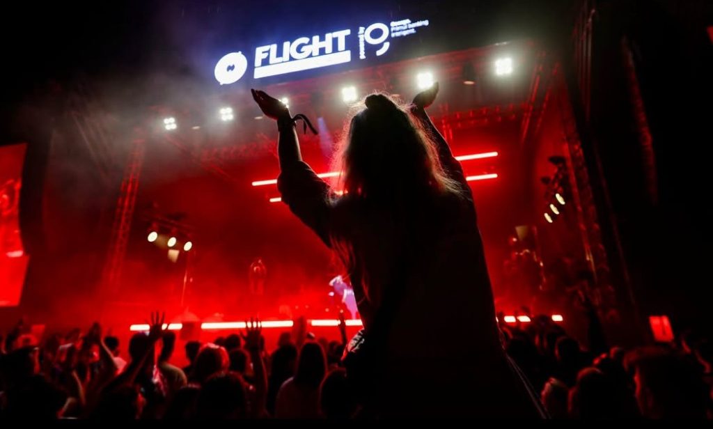 flight festival 2 - Sursa de Vest Concert crowd at night with a person on someone's shoulders raising arms, red stage lights, and a bright 'FLIGHT IO' sign above the stage.