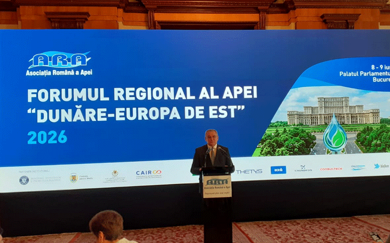 Speaker stands at a podium delivering a talk at a conference stage, in front of a large blue banner reading 'FORUMUL REGIONAL AL APEI Dunăre-Europa de Est 2026' with logos along the bottom edge of the screen and a building illustration on the right side.