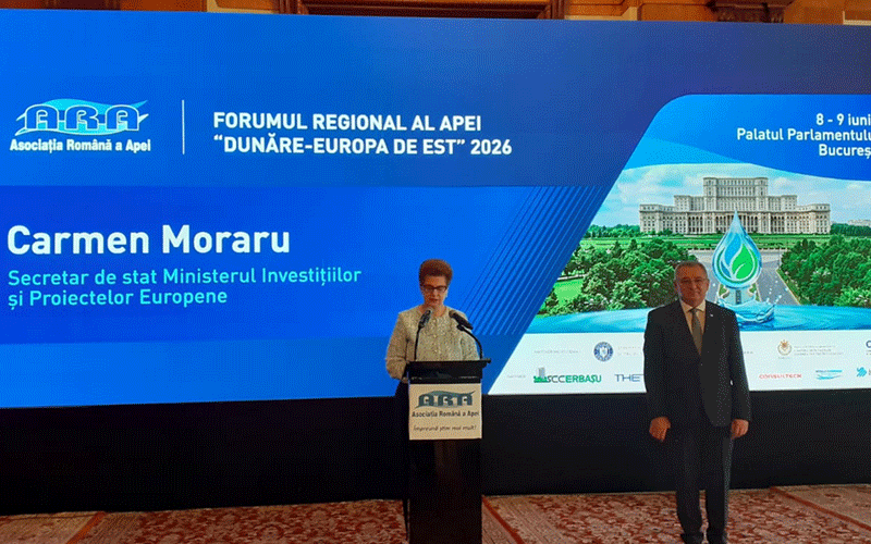forum-aquatim3 - Sursa de Vest Speaker at a podium delivering a speech on a blue conference stage with banners for the ‘Dunăre-Europa de Est 2026’ event and sponsors, Carmen Moraru shown on screen, a man in a suit stands nearby.