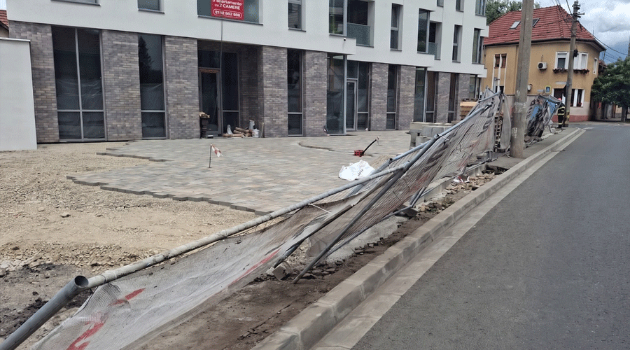 Leaning temporary construction fence along a curb beside a paved plaza in front of a modern building under construction.