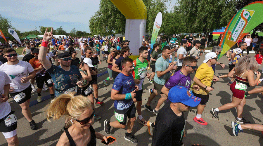 Crowd of runners at a lively outdoor race start, many wearing numbered bibs and sportswear under a bright sky with banners and inflatable arches ahead.