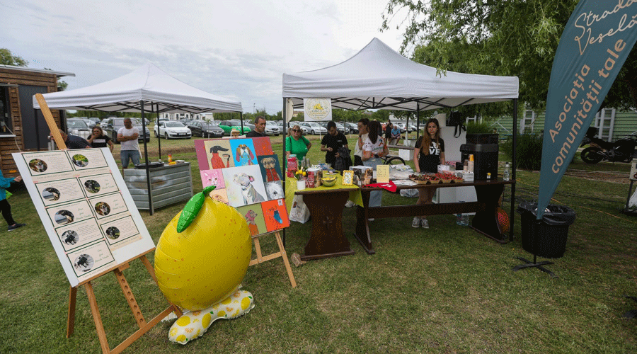 Outdoor community fair booth with volunteers behind a table of crafts and food under two white tents; a large yellow lemon sculpture on a stand nearby.