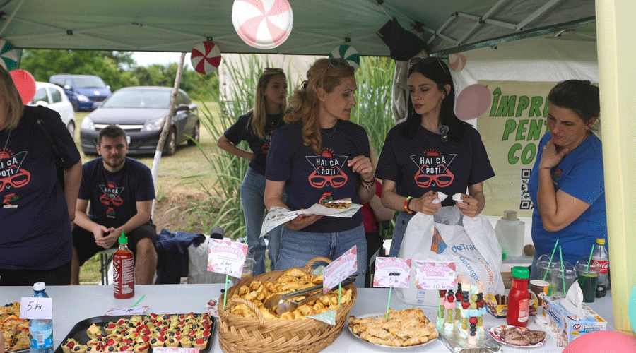 Group of volunteers in matching dark shirts behind a food stall at an outdoor event, serving and chatting over a table of pastries and snacks.