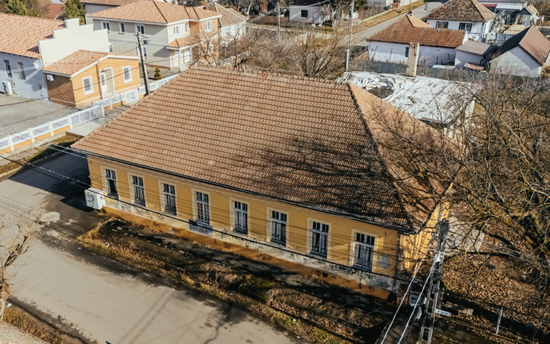 Aerial view of a yellow, single-story building with a red tile roof in a residential neighborhood, bordered by a street and trees.