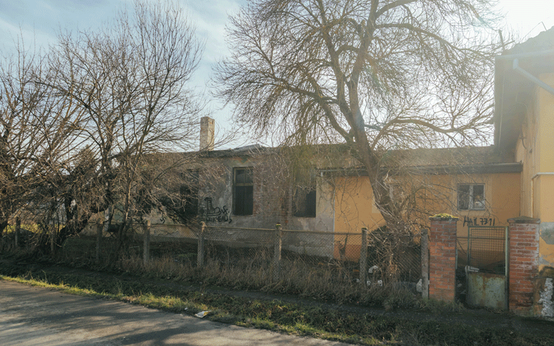 gradinita-plopi2 - Sursa de Vest Abandoned one-story house behind a wooden fence, graffiti on the walls, and bare trees along a quiet street.