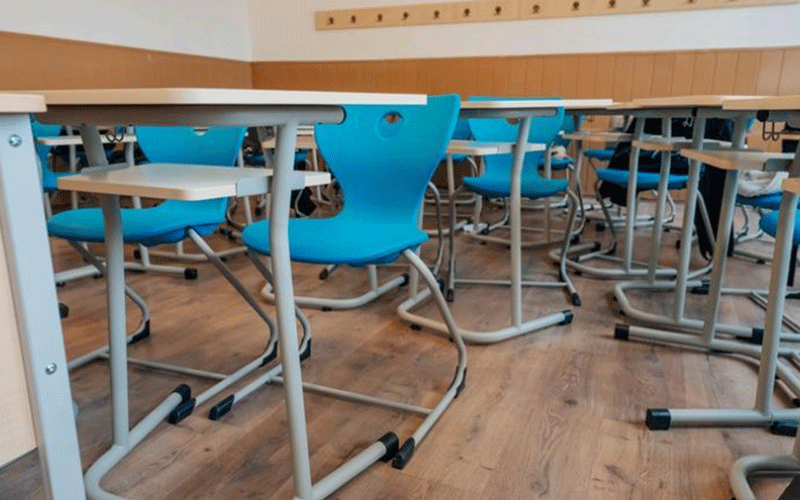 Empty classroom with blue plastic chairs tucked under light wooden desks, viewed from a low angle across a wood floor.