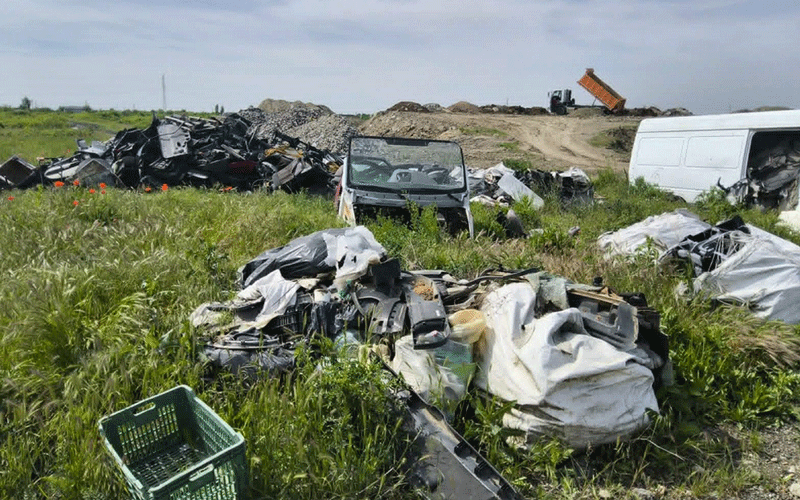 Scrap-yard scene: heaps of metal and debris in a grassy field, a small utility vehicle in front, and a white van in the background.