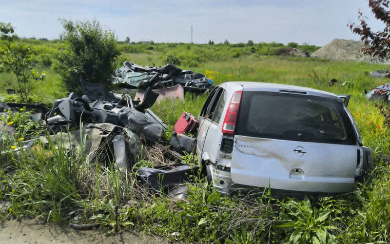 White station wagon wrecked in a grassy field with scattered car parts and debris around the vehicle