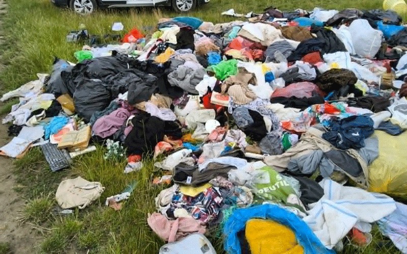 Large pile of assorted clothes scattered on grass beside a road, as if dumped illegally.