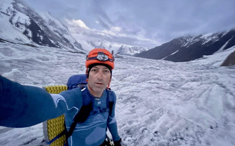 Man in blue jacket and orange helmet taking a selfie on a snowy glacier with mountains in the background, backpack on.