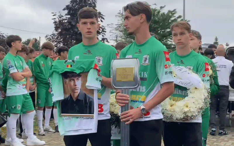 Group of young soccer players in green uniforms gather for a trophy ceremony; three front players hold a silver trophy and a framed portrait.