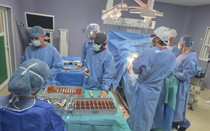 Surgical team in blue gowns and masks perform an operation around a patient on the operating table under overhead lights.