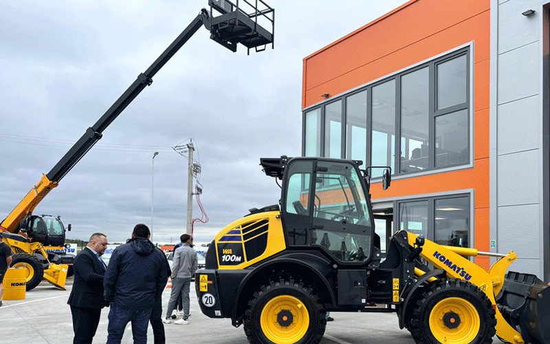 kuhn-romania2 - Sursa de Vest Yellow Komatsu front-end loader parked outside an orange-and-gray building, with a crane in the background and a small group of people nearby, suggesting a construction equipment event or demonstration.