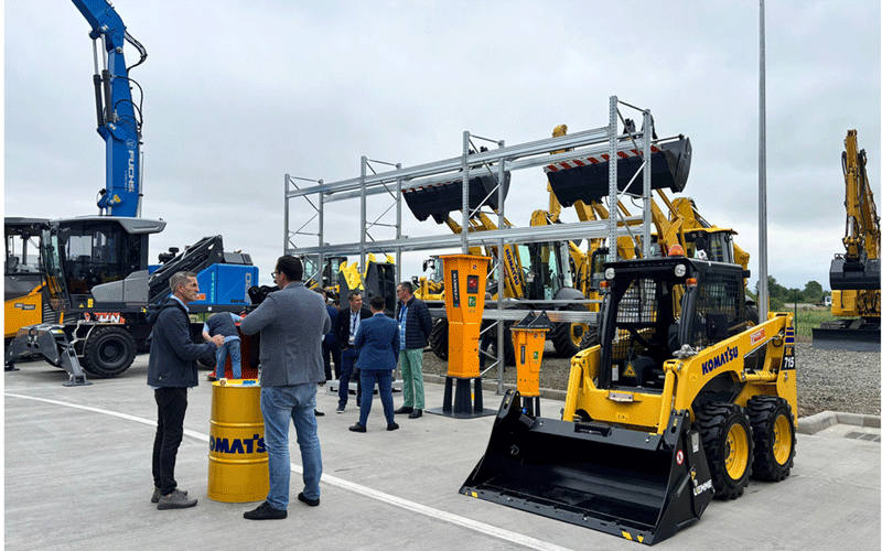 kuhn-romania4 - Sursa de Vest Group of people talking near a yellow Komatsu skid-steer loader at an outdoor construction equipment display with scaffolding and heavy machinery in the background.