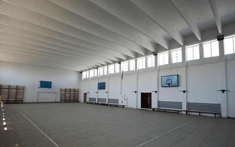 liceu-auto2 - Sursa de Vest Empty indoor gymnasium with multiple basketball hoops mounted on white walls, benches along the right wall, and high windows along the top letting in light.