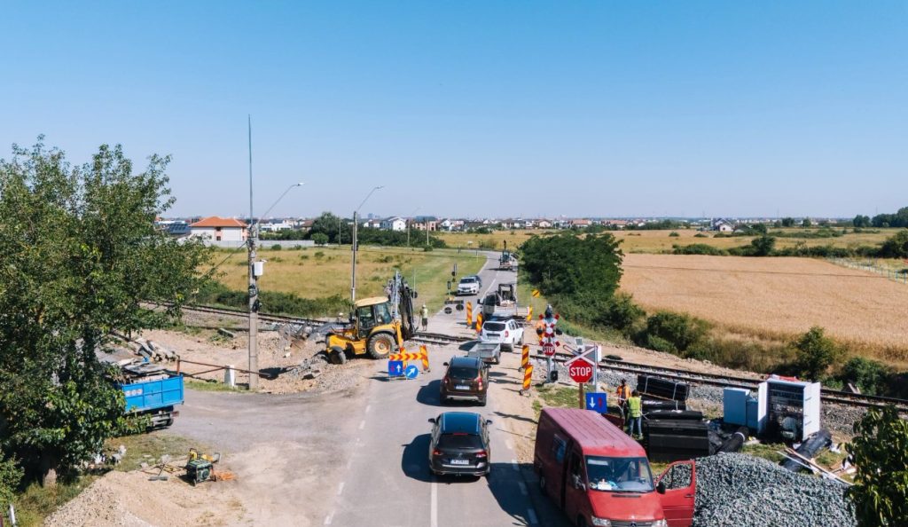 Road construction at a level crossing with a yellow excavator, barriers, and stopped cars on a rural road beneath a clear blue sky.