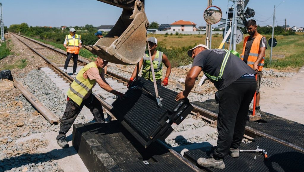Construction workers in safety vests install a black metal grate onto railroad tracks using a crane in a rural area.