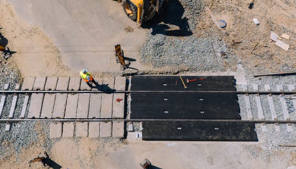 Construction workers lay railroad tracks; a worker in a high-visibility vest walks along concrete sleepers beside black modular track panels.