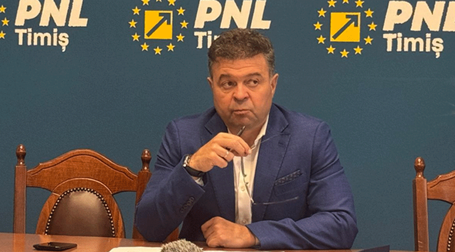 Man in a blue suit seated at a table during a press conference, holding a microphone with a PNL Timiș backdrop behind him.