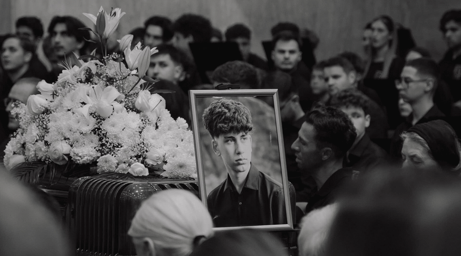 Framed portrait of a young man on a coffin surrounded by white flowers at a funeral with mourners in the background.