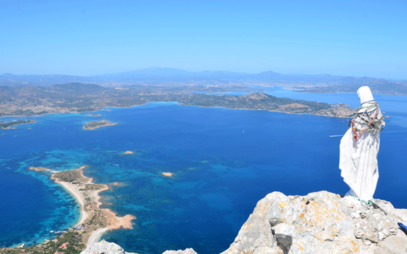 White-robed religious statue standing on a rocky cliff overlooking a turquoise sea and distant islands under a clear blue sky.