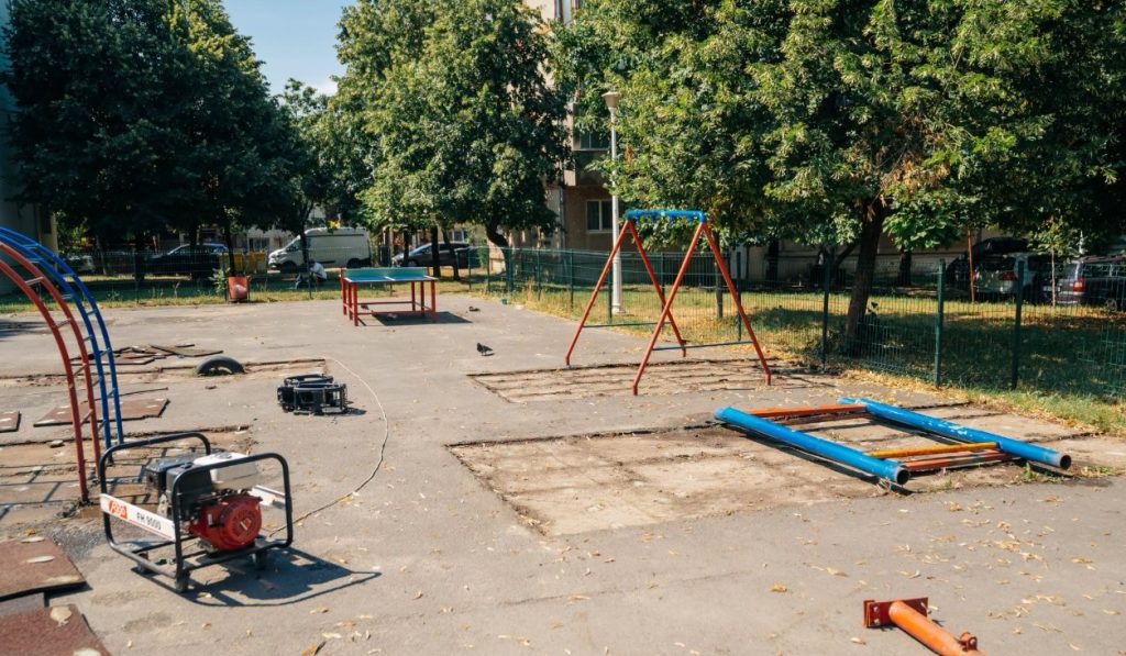 Disassembled playground in a fenced park: swing frame, blue bars, scattered pipes, and a portable generator on the left edge of the scene.