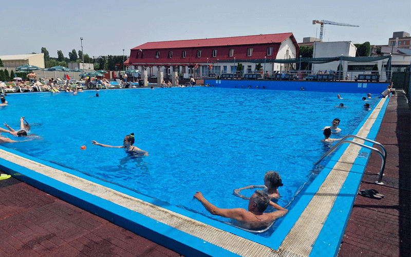 Outdoor public swimming pool scene with people swimming and sunbathing around a bright blue pool, a red-roofed building in the background.