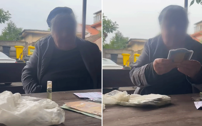 Person with blurred face sits at an outdoor table, looking at a smartphone; cash, tissues, and a small bottle on the table are visible. Alternative wording: Outdoor scene with a person using a phone; items on the table include money, tissues, and a tiny bottle.