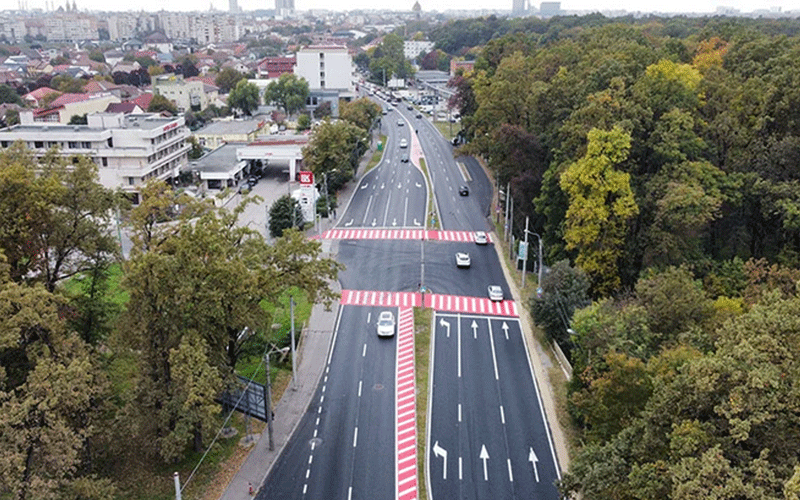 Aerial view of a multi-lane road with red-and-white striped crosswalk barriers and surrounding trees and buildings far in the distance.