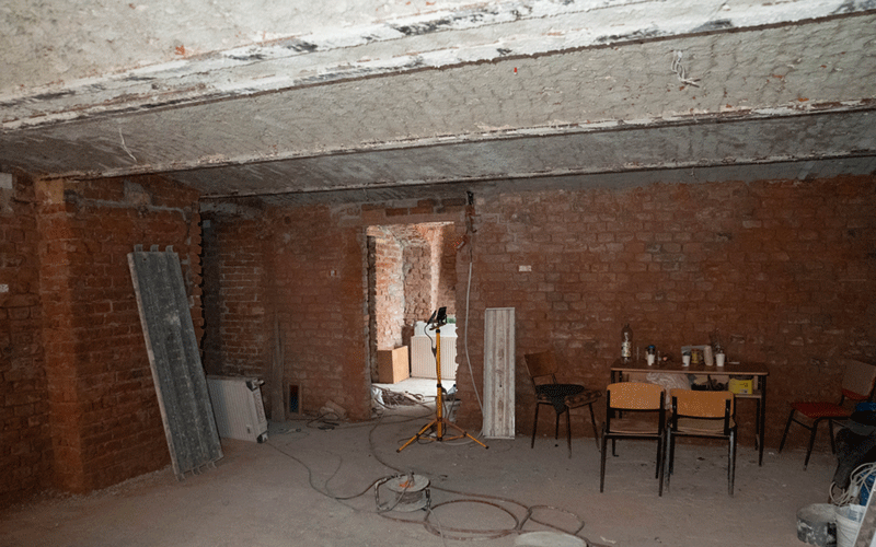 reparatii-scoli - Sursa de Vest Unfinished brick-walled basement with construction equipment and a table of chairs set against the wall under a concrete beam, dusty floor scattered with cables and tools.