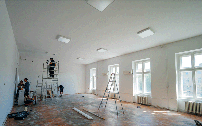 reparatii-scoli2 - Sursa de Vest Construction workers renovate a large sunlit room with scaffolding, ladders, and tools on the floor during a renovation project, with tall windows in the background and bright white walls.